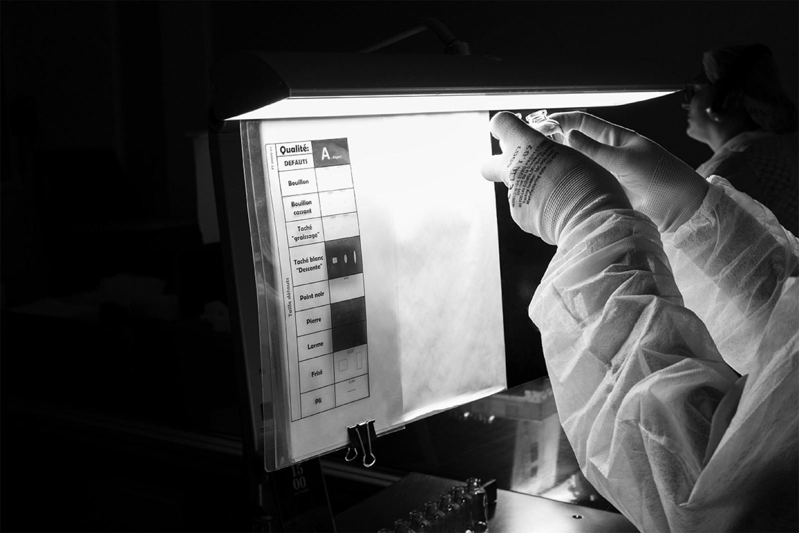 Visual inspection of a bottle held up to a light panel, in cleanroom garments